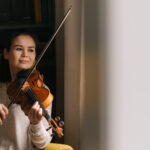 Fiddler plays tender music on a violin in her home near the window.